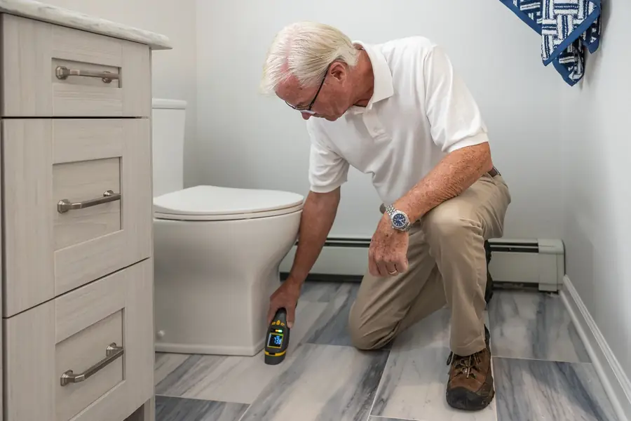 Man kneeling beside a toilet to check the bathroom floor with a handheld meter